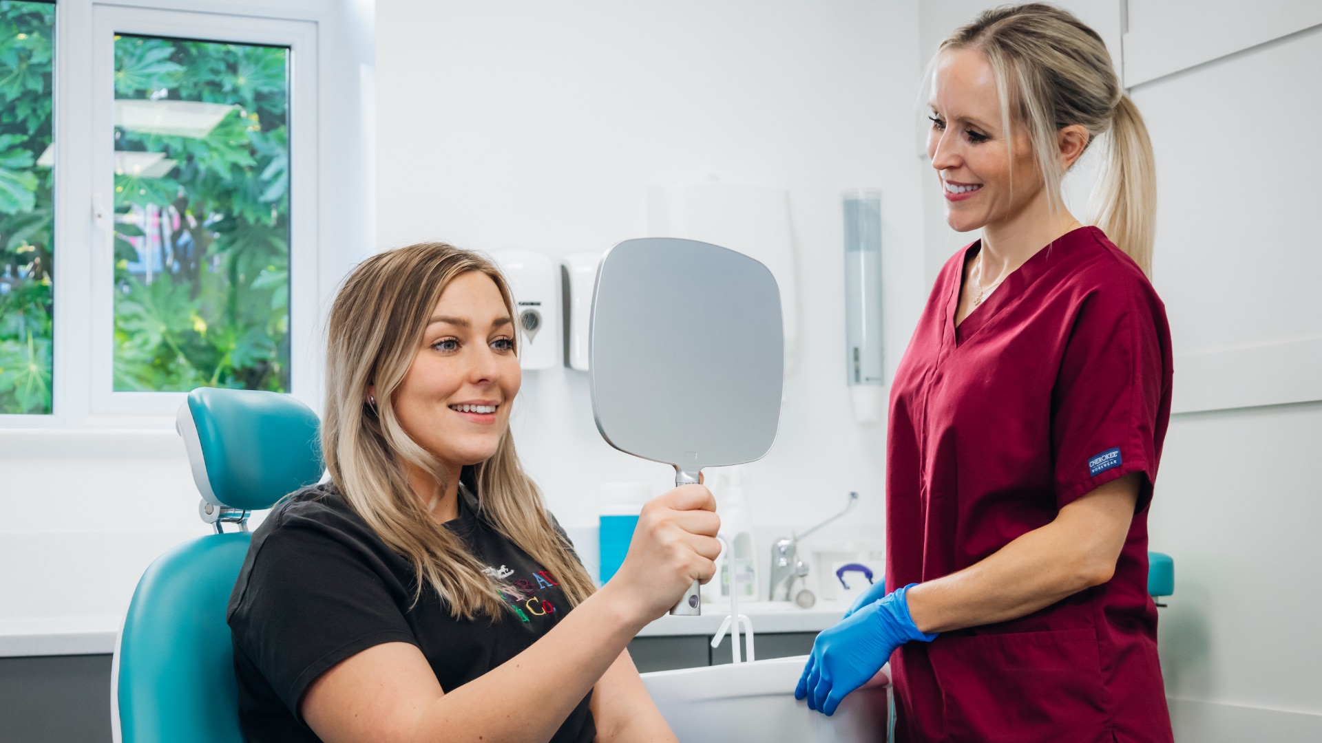 A blonde female in the treatment chair looking at herself in a grey handheld mirror as Dr Hannah Wilson, a blonde clinician smiles at the patient