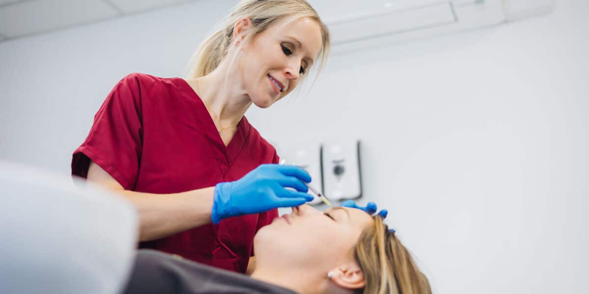 Dr Hannah Wilson, a blonde clinician wearing maroon scrubs and blue latex gloves injecting a syringe of filler into a female patients forehead