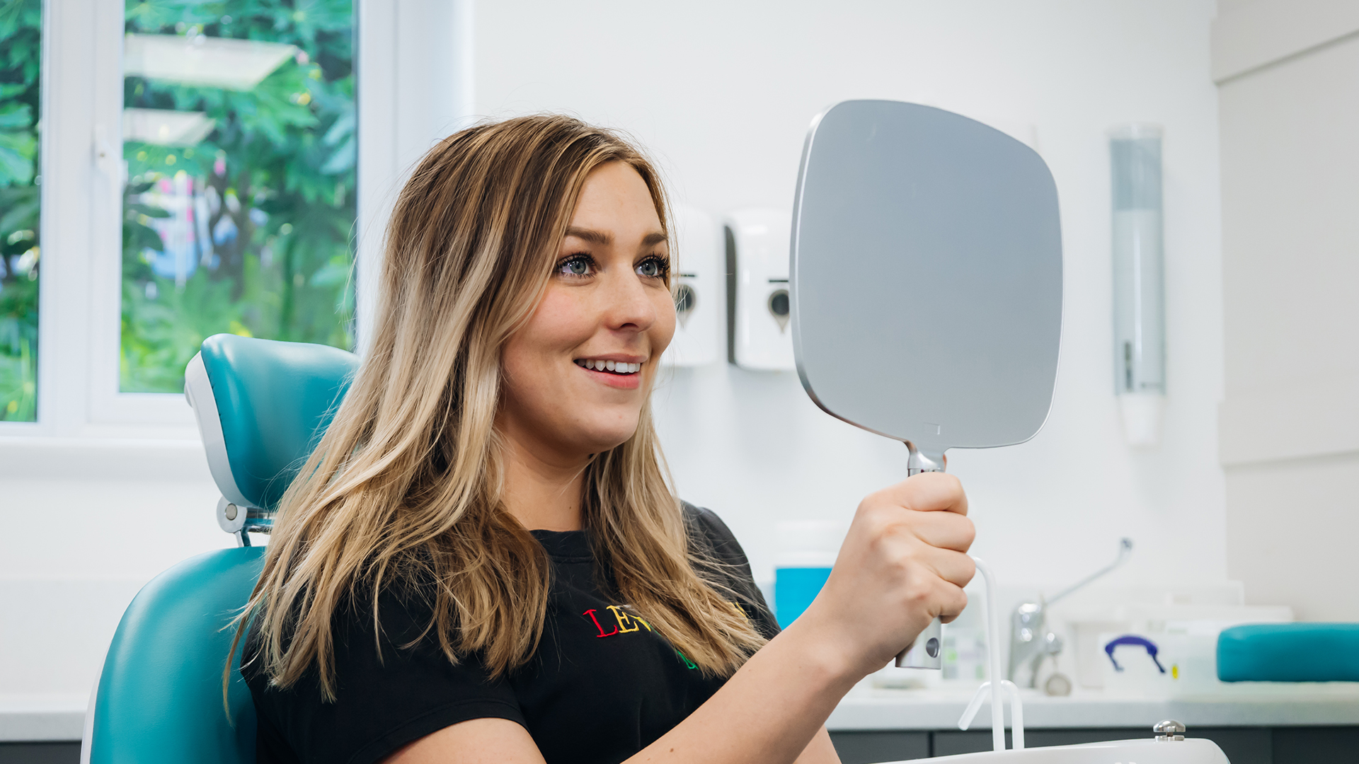 A female patient with blonde hair looking into a handheld mirror and smiling