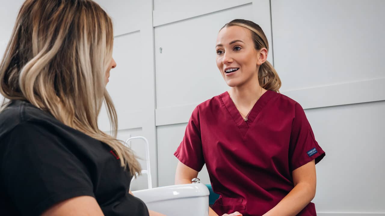 A female clinician wearing maroon scrubs talking to a patient and smiling