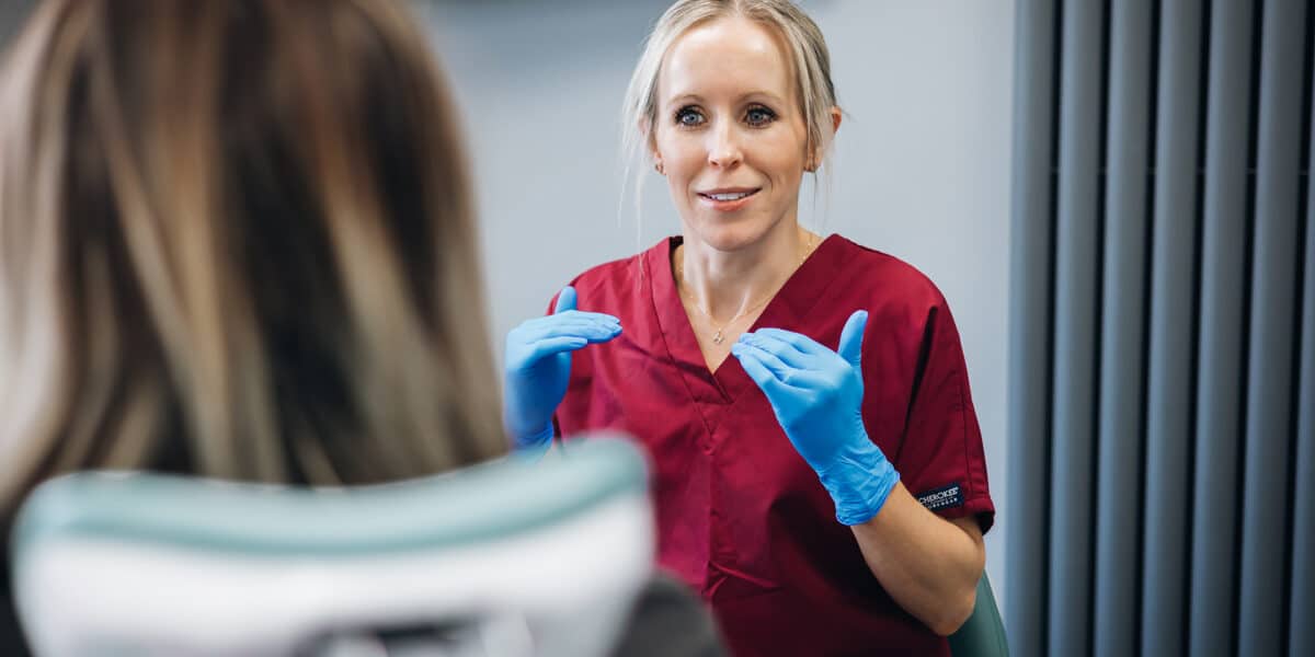 Dr Hannah Wilson, a blonde clinician wearing maroon scrubs and blue latex gloves smiling while gesturing with her hands