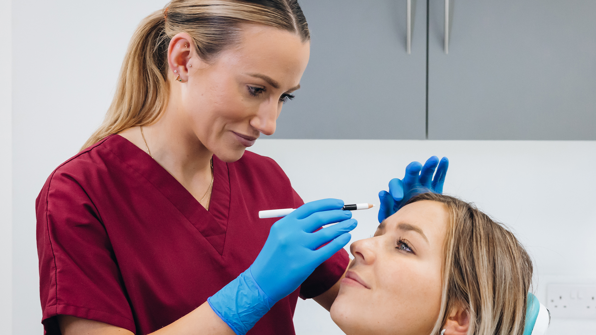 A female clinician wearing maroon scrubs and blue latex gloves using a white pencil on a female patients face